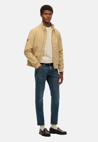 Beige letvægtsjakke med ståkrave, lynlås-lukning, parret med en cremefarvet sweater, blå jeans og brune loafers.
