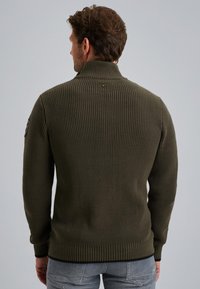 Olijfgroen brei-sweater met een hoge kraag, ribbelstructuur en een zwarte zoom. Heeft een patch op de linker mouw.