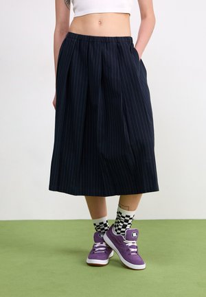 Jupe midi rayée marine foncé avec taille élastique et poches latérales, associée à des baskets violettes et des chaussettes à carreaux sur un sol vert.
