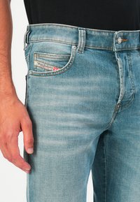 Primer plano de una persona que lleva puestos unos vaqueros de mezclilla azul claros con una pequeña etiqueta en el bolsillo delantero y una mano descansando al lado del muslo.