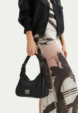 Kobieta w kurtce bomberce i wzorzystej spódnicy trzymająca czarną skórzaną torebkę z metalowym logo G-Star Originals.