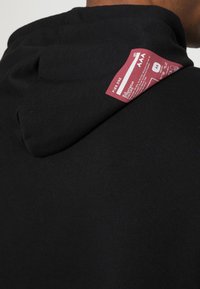 Felpa nera con cappuccio e texture a coste; presenta un'etichetta rossa nella parte posteriore, vicino al collo, che mostra le istruzioni per la cura e il nome del marchio.