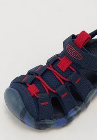 Sandalia azul marino con acentos rojos, correas de malla y suela de goma. Presenta una correa ajustable y una plantilla texturizada para mayor agarre.