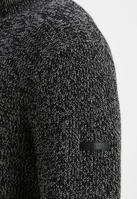 Gestrickter Pullover in Schwarz und Weiß mit einem strukturierten Muster. Verfügt über ein schwarzes Markenzeichen auf dem linken Ärmel.