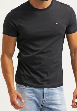 Mand iført en tætsiddende sort t-shirt med rund hals og en lille rød, hvid og blå logo på venstre bryst samt lyseblå jeans.