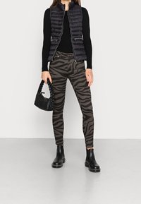 Frau trägt eine schwarze gesteppten Weste, ein schwarzes Langarmoberteil, zebra-musterte Hose, schwarze Stiefeletten und hält eine geflochtene schwarze Handtasche.