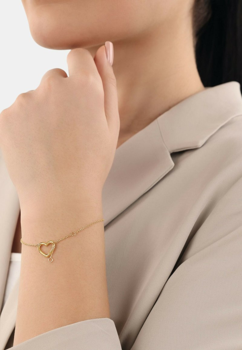 Bracciale d'oro con un ciondolo a forma di cuore, catena fine e un delicato gancetto. La mano del modello è appoggiata su un morbido tessuto di colore neutro.