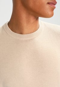 Beiger Strickpullover mit Rundhalsausschnitt, glatter Textur und geripptem Besatz am Kragen, welcher eine taillierte Silhouette betont.