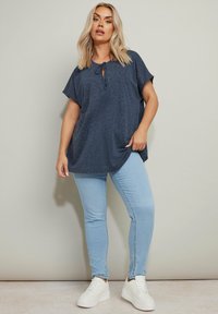 Maglietta oversized blu navy con maniche corte, con dettaglio a cordoncino sul collo, abbinata a jeans slim-fit azzurri e sneakers bianche.