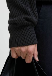 Puño de suéter negro de canalé alrededor de una mano que sostiene un bolso de cuero negro. La tela es texturada y ajustada, con un acabado suave en el bolso.