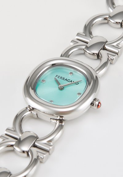 Reloj plateado con esfera cuadrada, dial turquesa y brazalete metálico de eslabones. Destaca una piedra preciosa roja en la corona y marcadores de hora minimalistas.