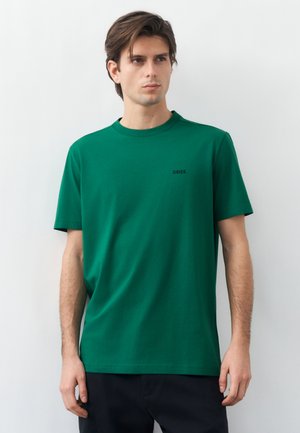 Νεαρός με καστανά μαλλιά που φοράει πράσινο κοντομάνικο μπλουζάκι BOSS και μαύρο παντελόνι, στέκεται μπροστά σε απλό φωτεινό φόντο.