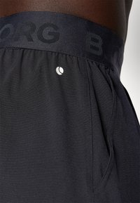 Calções atléticos pretos com uma cintura texturizada, apresentando "BORG" em impressão escura. Detalhe de logotipo reflexivo na lateral. Tecido suave.