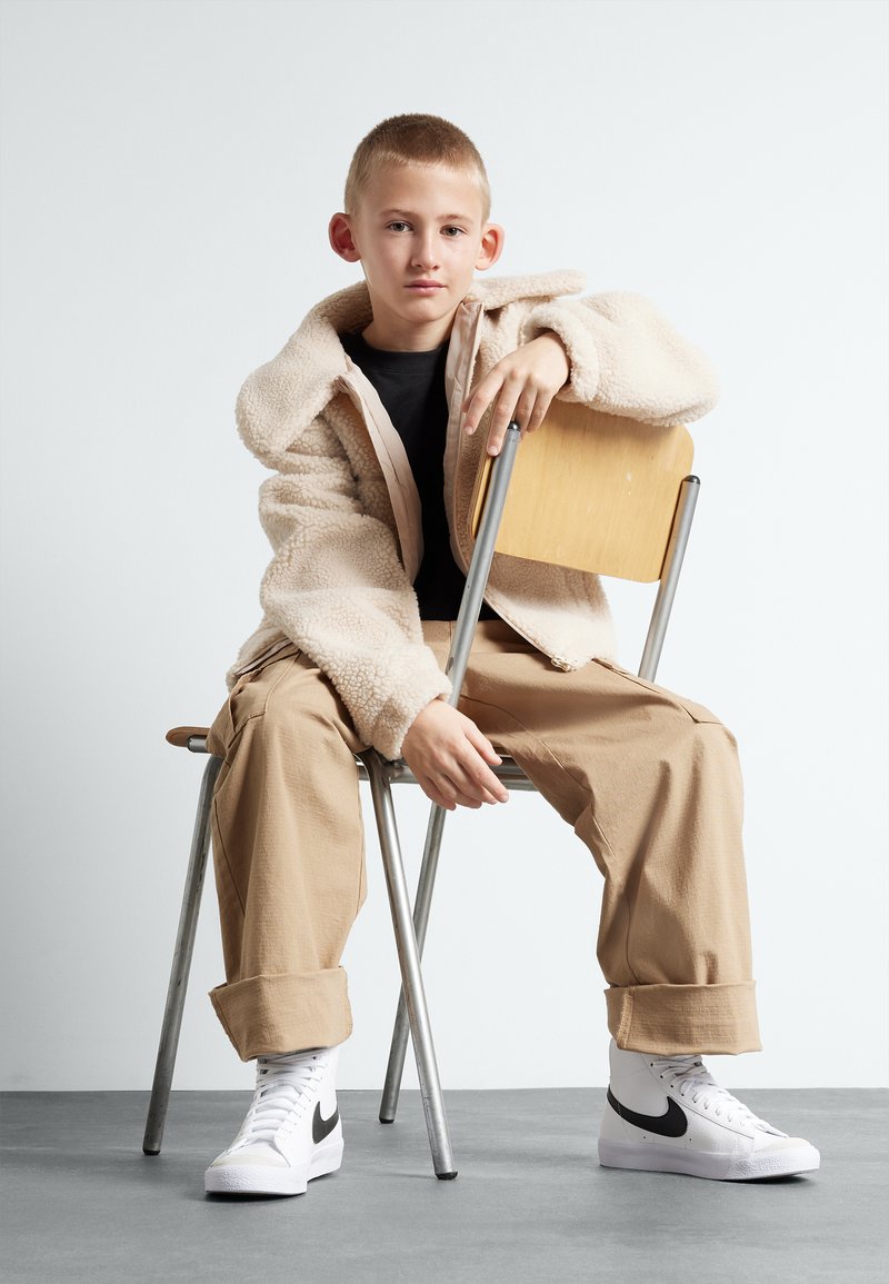 Chaqueta de forro polar color crema, camisa negra, pantalones beige y zapatillas blancas con acentos negros. Sentado en una silla de metal, fondo neutro.