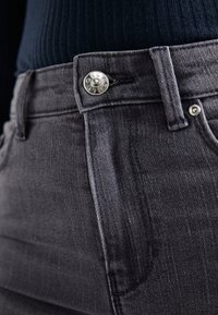 Temno sivi jeans iz denima z metalnim gumbom, oblečenimi v pasu in s stranskimi šivi. Material ima gladko teksturo.