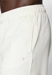 Cremfarbene Sweatpants aus weichem Stoff, mit verstellbarem Bund, Seitentasche und dezentem gesticktem Logo auf dem Oberschenkel.