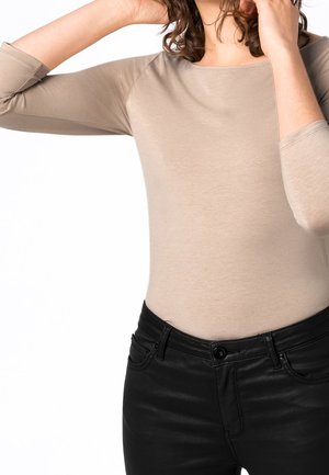 Persoon draagt een beige top met lange mouwen en een zwarte broek, met één arm omhoog, haar haar aanpassend, tegen een witte achtergrond.