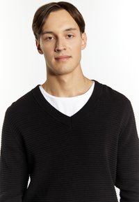 Pullover negru texturat cu un decolteu în V, purtat peste un tricou alb. Puloverul are un material ribbat și mâneci lungi.