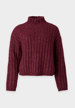 Jeune personne debout, mains derrière le dos, portant un pull en maille bordeaux, un pantalon ample beige et des chaussures beiges sur un fond uni.