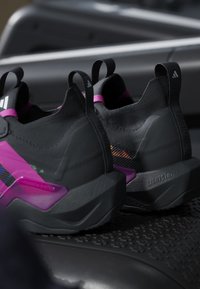 Μαύρα και μωβ αθλητικά παπούτσια Adidas με θηλιές, υφασμένες σόλες και μαλακά ενδιάμεσα πέλματα τοποθετημένα σε λαστιχένιο χαλάκι.
