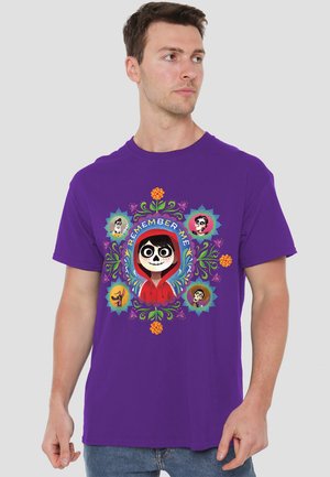 Mann in lila Hemd mit buntem "Remember Me"-Design, das eine Figur in einer roten Kapuze zeigt, umgeben von floralen Mustern und kleinen Figuren.