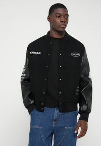 Fekete bomber dzseki bőr ujjal, hímzett logókkal és gombokkal. Fekete pulóverrel és farmerekkel kombinálva.