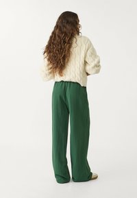 Vrouw met lang, krullend haar, gekleed in een crèmekleurige kabelgebreide trui en groene wijde broek, staand met haar rug naar de camera op een witte achtergrond.