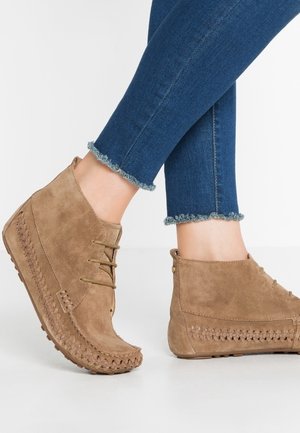 Bottines en daim beige avec coutures tressées et lacets, portées avec un jean skinny bleu à ourlet effiloché, jambes croisées.