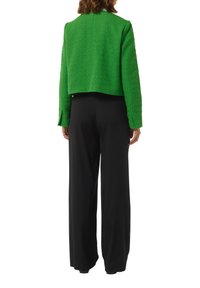 Groene cropped jas van kwetsbaar stof, met een ronde halslijn en lange mouwen; gecombineerd met zwarte wijde broek.