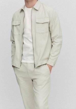 Mann trägt eine hellbeige Jacke mit Reißverschluss und Brusttaschen, weißes T-Shirt und passende beige Hose, Hände in den Taschen, steht vor einfachem Hintergrund.