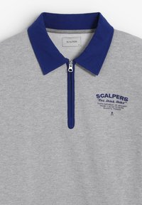Grijze polo met een blauwe kraag, half-rits aan de voorkant en een stoffen label. Kenmerkt zich door blauwe geborduurde tekst die de locatie en merknaam vermeldt.