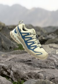 Hellbeiger Sportschuh mit blauen Akzenten, Mesh-Obermaterial, Schnürsystem und strukturierter Sohle, entworfen für Outdoor-Aktivitäten.
