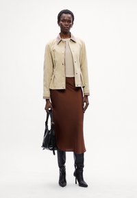 Casaco bege com gola castanha, blusa creme, saia midi castanha e botas de cano alto pretas. Segura uma mala preta. Texturas suaves e linhas limpas.