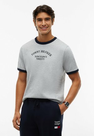 Mand smiler iført en grå Tommy Hilfiger t-shirt med sort kant og marineblå bukser med Tommy Hilfiger-logo.