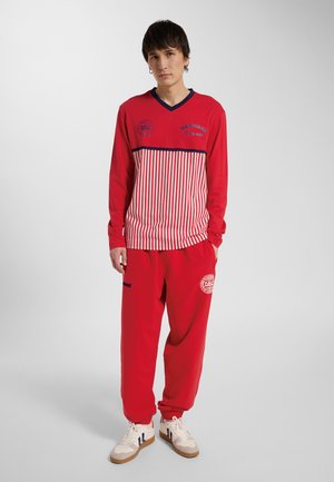 Ung mand stående med hænderne i lommerne iført en rød og hvid stribet langærmet top og røde joggingbukser med hvide sneakers.