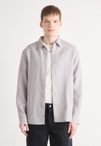Lichtgrijze overhemden van een textuurachtige stof, voorzien van een klassieke kraag en metalen knopen, gecombineerd met een wit t-shirt en donkere jeans.