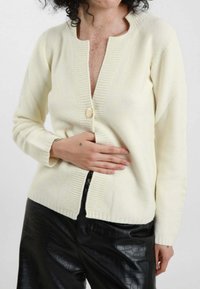 Cardigan lavorato a maglia crema con scollo rotondo, polsini a coste e un singolo bottone dorato. Indossato sopra pantaloni neri con texture.