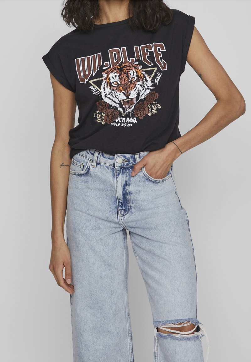 Personne portant un débardeur noir avec un motif tigre rugissant et texte, associé à un jean bleu clair déchiré.