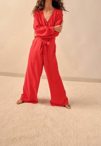 Combinaison rouge à manches longues avec un col en V et une fermeture éclair à l'avant. Dotée d'une ceinture à nouer et de jambes larges, confectionnée dans un tissu lisse. Portée avec des sandales à brides.