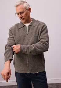 Grijze wollen zip-up cardigan met een kraag, geribde manchetten en een relaxte pasvorm, gedragen over een wit shirt, gecombineerd met donkerblauwe broek.