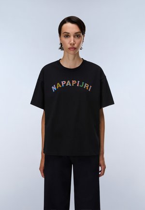 Femme aux cheveux courts et foncés porte un t-shirt noir oversize avec un texte coloré "NAPAPIJRI", debout devant un fond clair uni.
