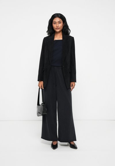 Blazer en velours côtelé noir sur un top navy, associé à un pantalon noir ample et des escarpins noirs à bout pointu. Le mannequin tient un petit sac à main noir.