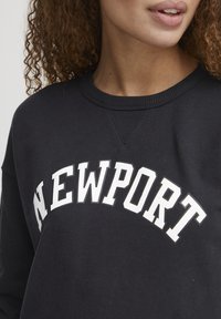 Felpa nera con lettering bianco "NEWPORT". Presenta un collo a giro costolato e una texture morbida. Inquadratura ravvicinata della parte superiore del corpo.
