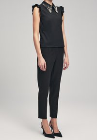 Top nero senza maniche con spalle arricciate, colletto decorato con accenti simili a perle, abbinato a pantaloni neri slim e tacchi.