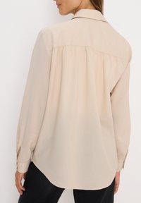 Chemise beige clair à manches longues dotée d'un col classique, de détails plissés dans le dos et de poignets à boutons. Tissu lisse et doux.