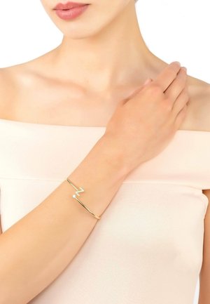 Gold bracelet featuring a zigzag design with a small gemstone accent. Smooth, shiny texture on a wrist against a light-colored top.