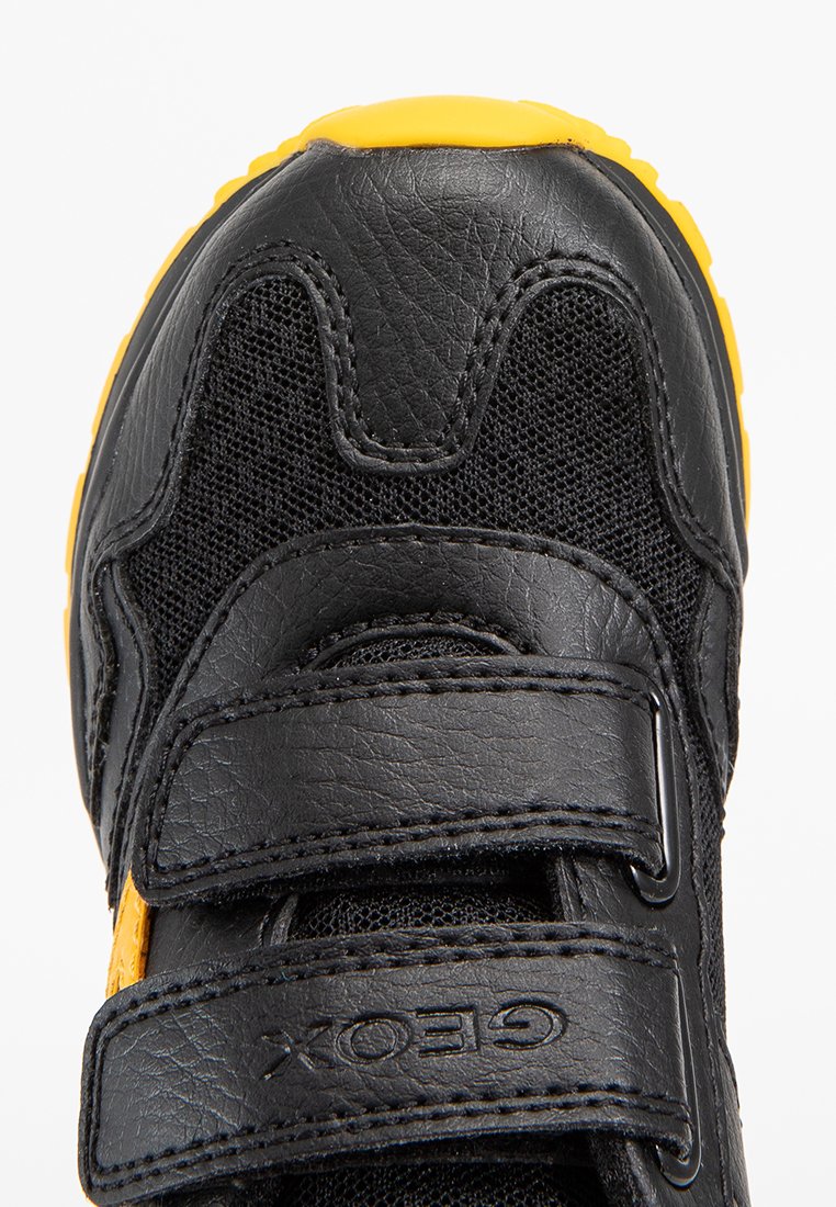 Chaussure noire en matériaux en maille et cuir, dotée de deux larges bandes Velcro. La semelle est jaune, avec un motif texturé pour une meilleure adhérence.