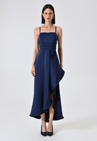 Marineblauwe satijnen asymmetrische jurk met dunne bandjes, gerimpelde taille en een bloemaccent. De zoom valt elegant met een zijsplit. Zwarte hakken.