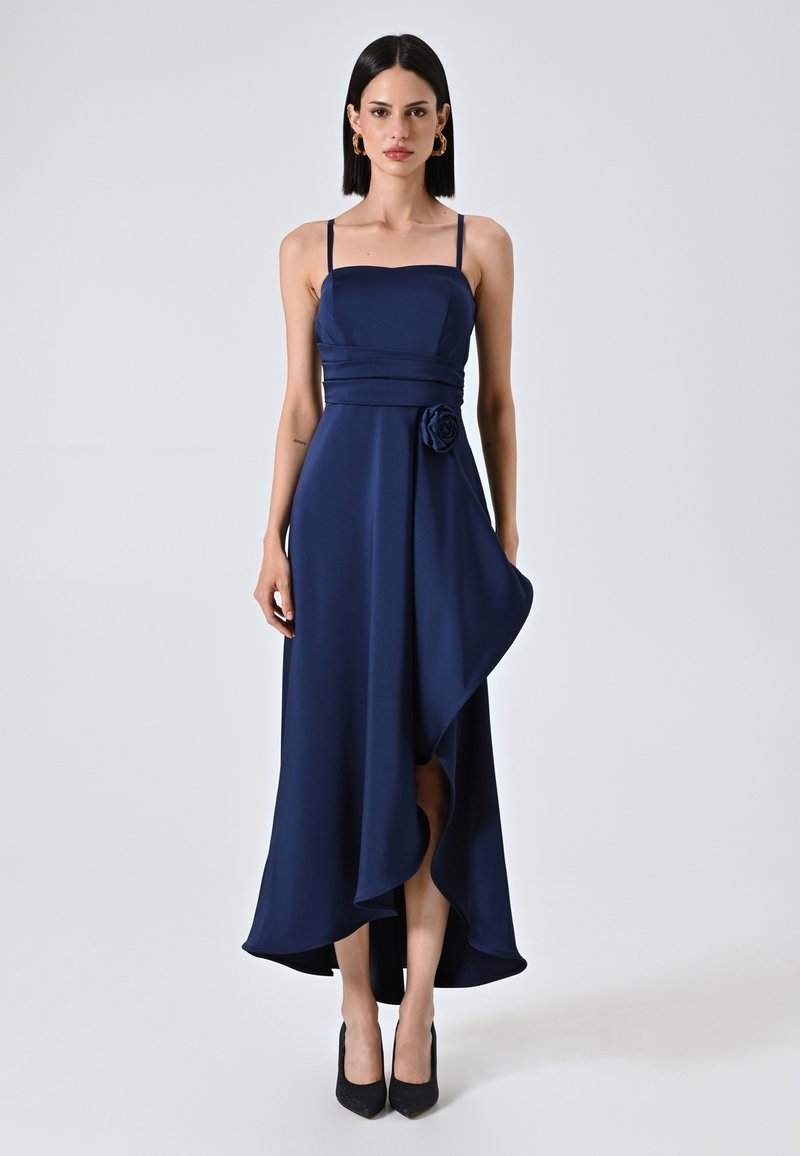 Marineblauwe satijnen asymmetrische jurk met dunne bandjes, gerimpelde taille en een bloemaccent. De zoom valt elegant met een zijsplit. Zwarte hakken.