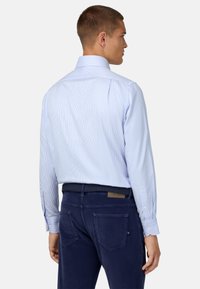 Gaiši zils svītrots krekliņš ar garām piedurknēm, pogājamiem aprocēm un izkliedētu apkakli, uzvilkts virs tumši ziliem biksēm. Aizmugures skats parāda krekliņa pielāgoto piegriezumu.